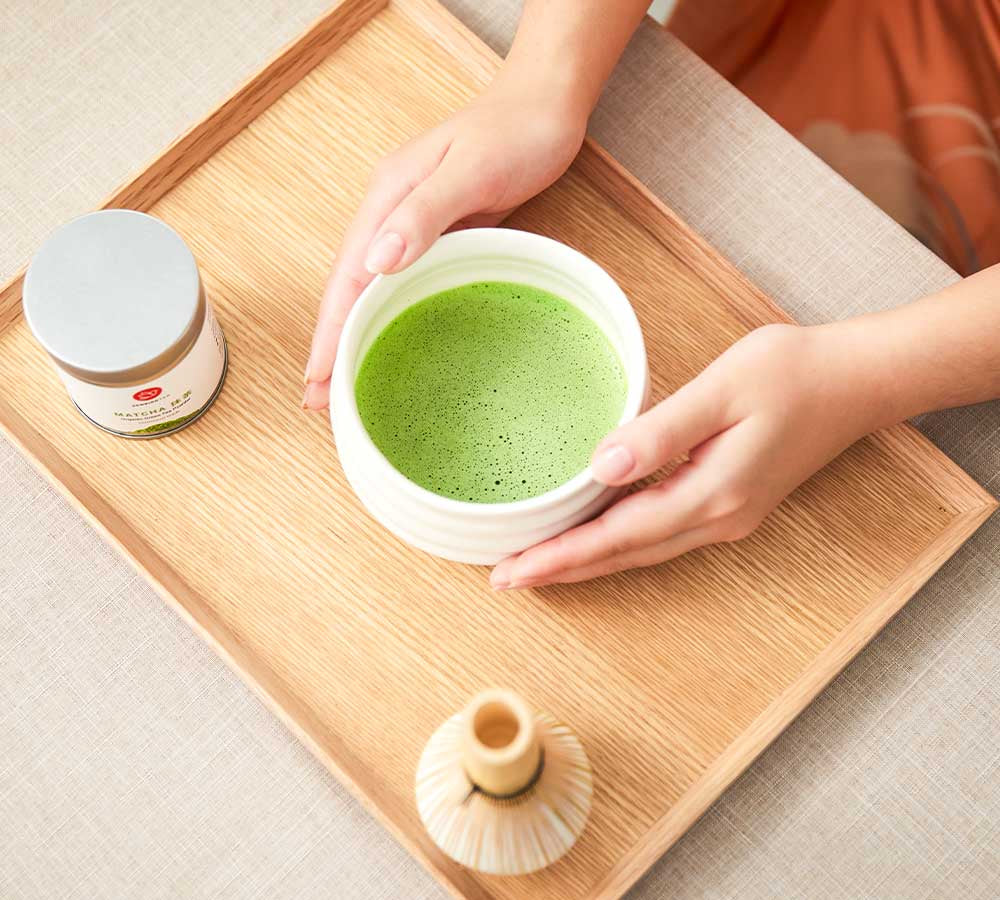 Hands holding a bowl of matcha beside a Senbird tea tin and bamboo whisk